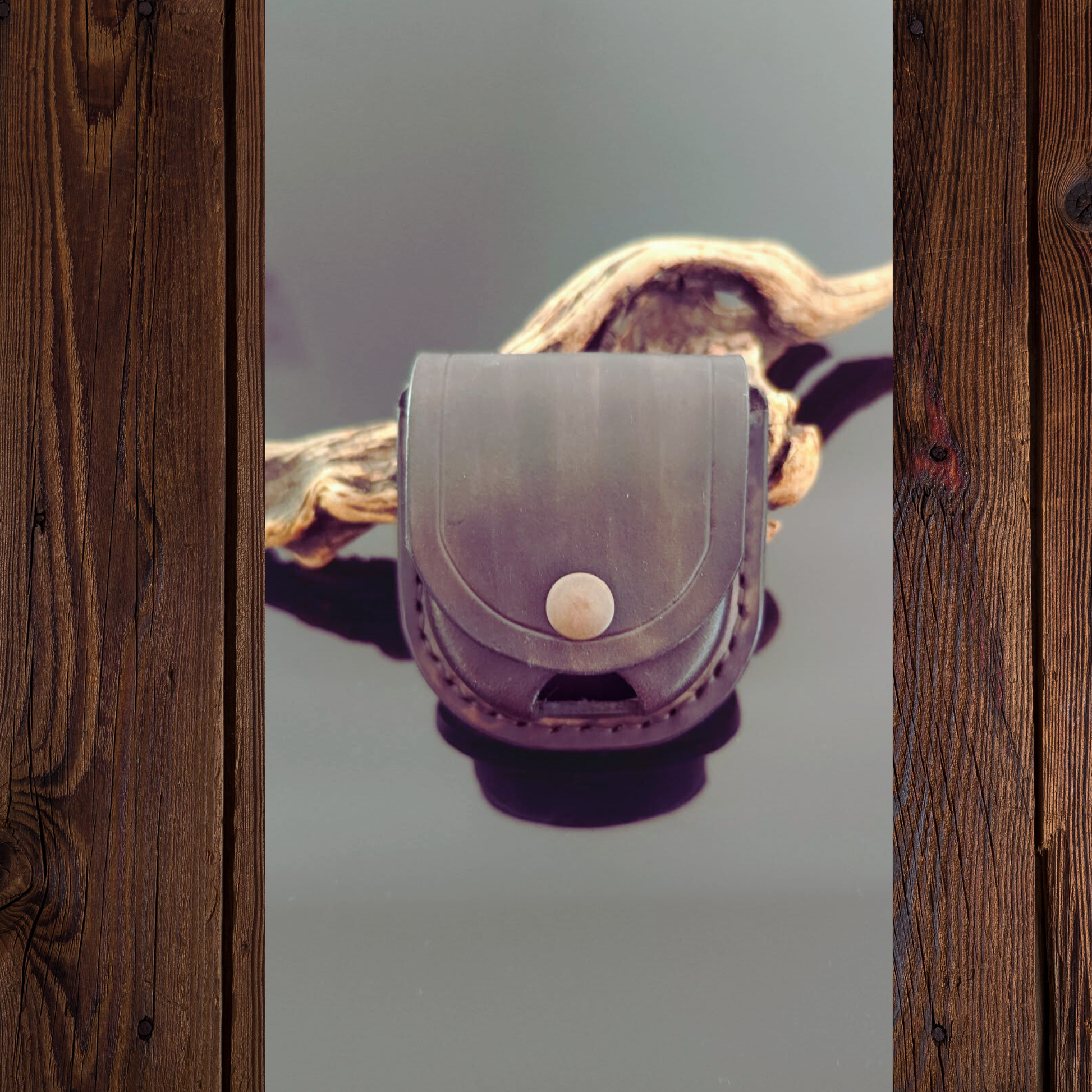 A small leather pouch or case with a flap closure and a single round button, shown against a neutral gray background with driftwood behind it. The image is framed by dark brown wooden boards on both sides.